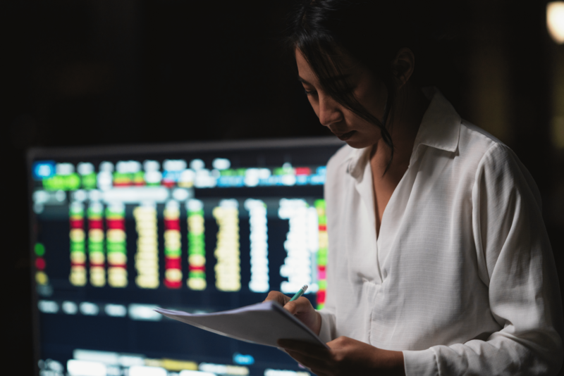 Woman with piece of paper and pencil in front of computers