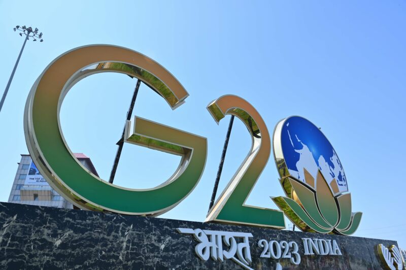 Ein gold umrandeter Schriftzug mit den Lettern "G20", der aus den Farben grün und orange besteht, steht neben dem Symbol einer Erde und einer Lotusblüte über dem Schriftzug "India 2023".