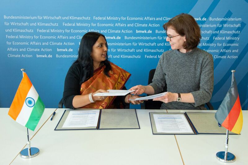 Zwei einander zugewandte Frauen tauschen ein Dokument aus. Im Vordergrund befinden sich eine deutsche und eine indische Flagge.