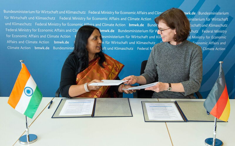 Two women facing each other exchange a document. In the foreground are a German and an Indian flag.